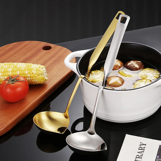 Two spoons, one gold and one silver, in a pot with food on a dark surface with a wooden board and vegetables.